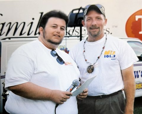 Two people standing outdoors with a microphone and notes, in front of a vehicle.