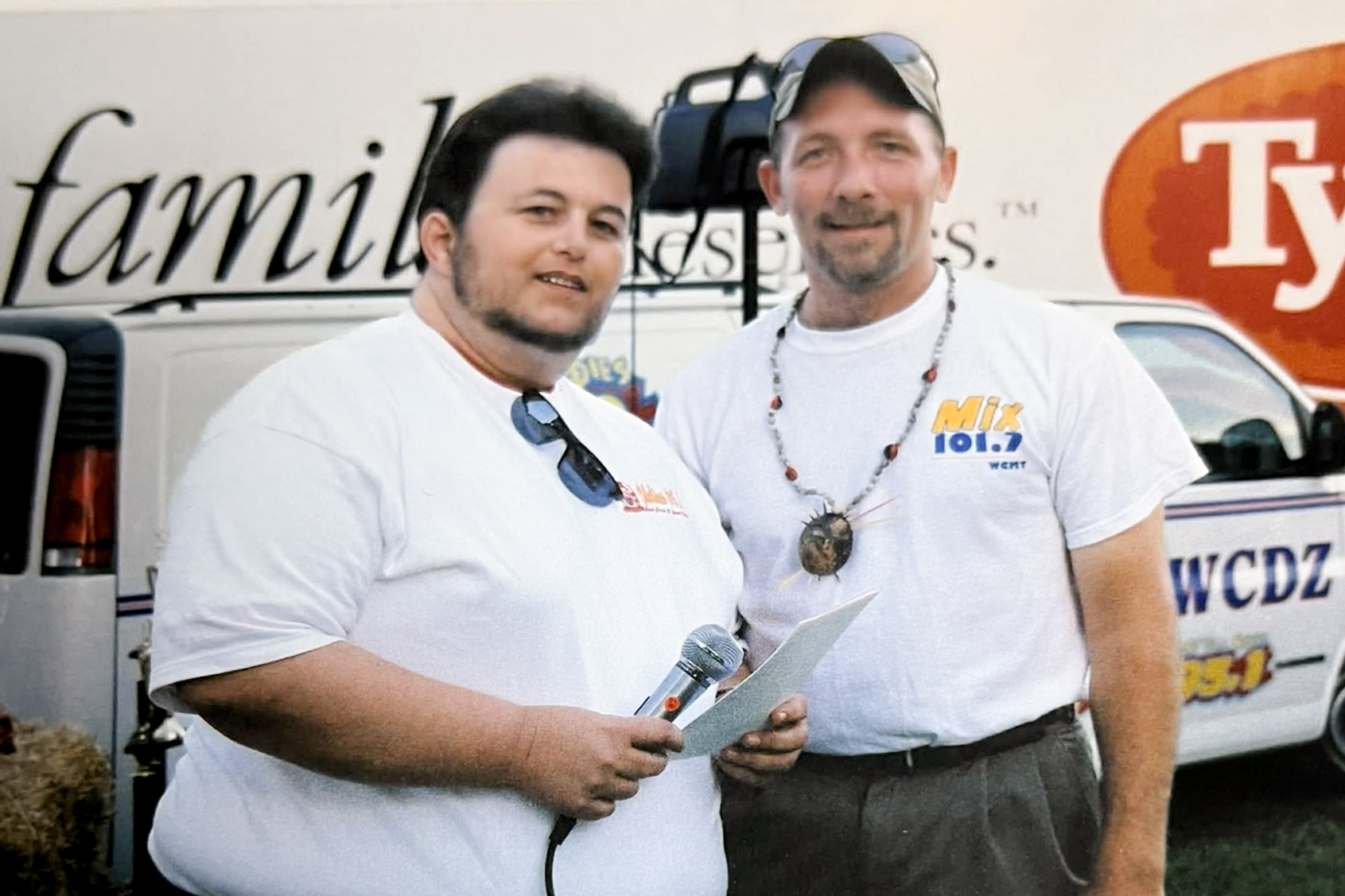 Two people standing outdoors with a microphone and notes, in front of a vehicle.