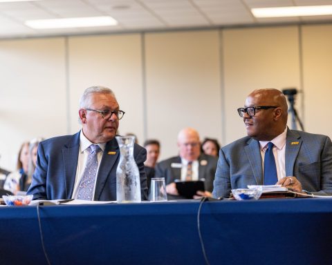 Two men in suits sitting at a table in a meeting setting with others in the background.