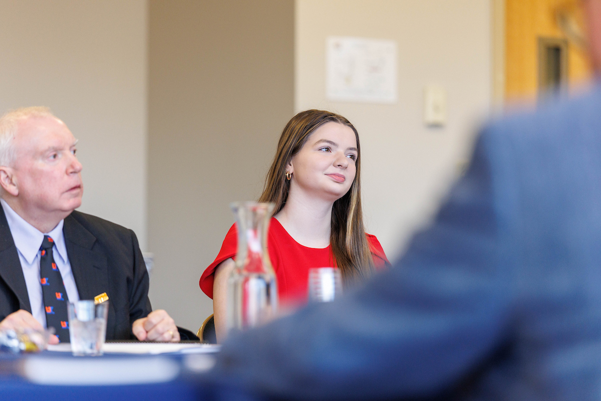 Two people seated at a table in a meeting room, with a glass pitcher and papers in front of them.