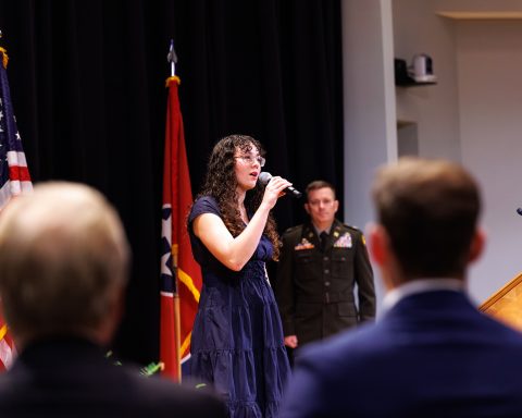 A woman sings into a microphone at a formal event, with a man in a military uniform behind her and flags to the left.