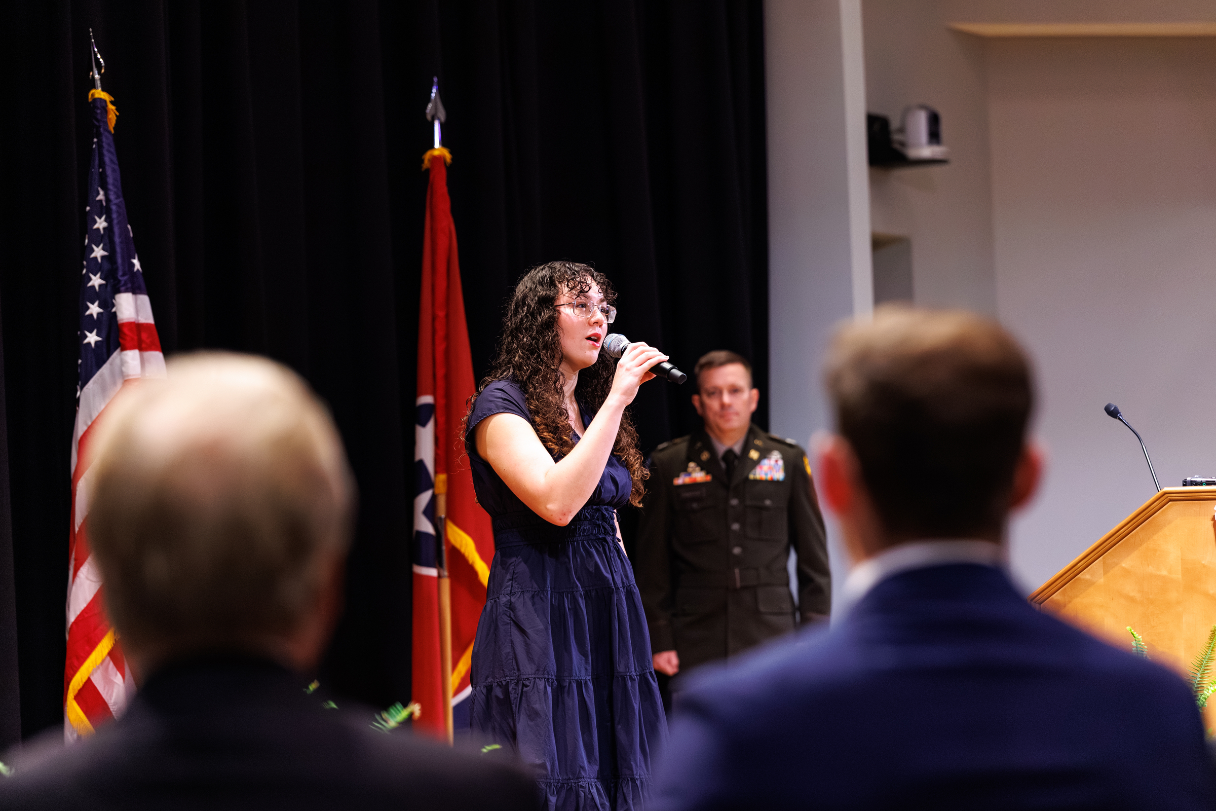 A woman sings into a microphone at a formal event, with a man in a military uniform behind her and flags to the left.