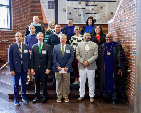 A group of people standing on an indoor staircase with a brick wall and plaques in the background.