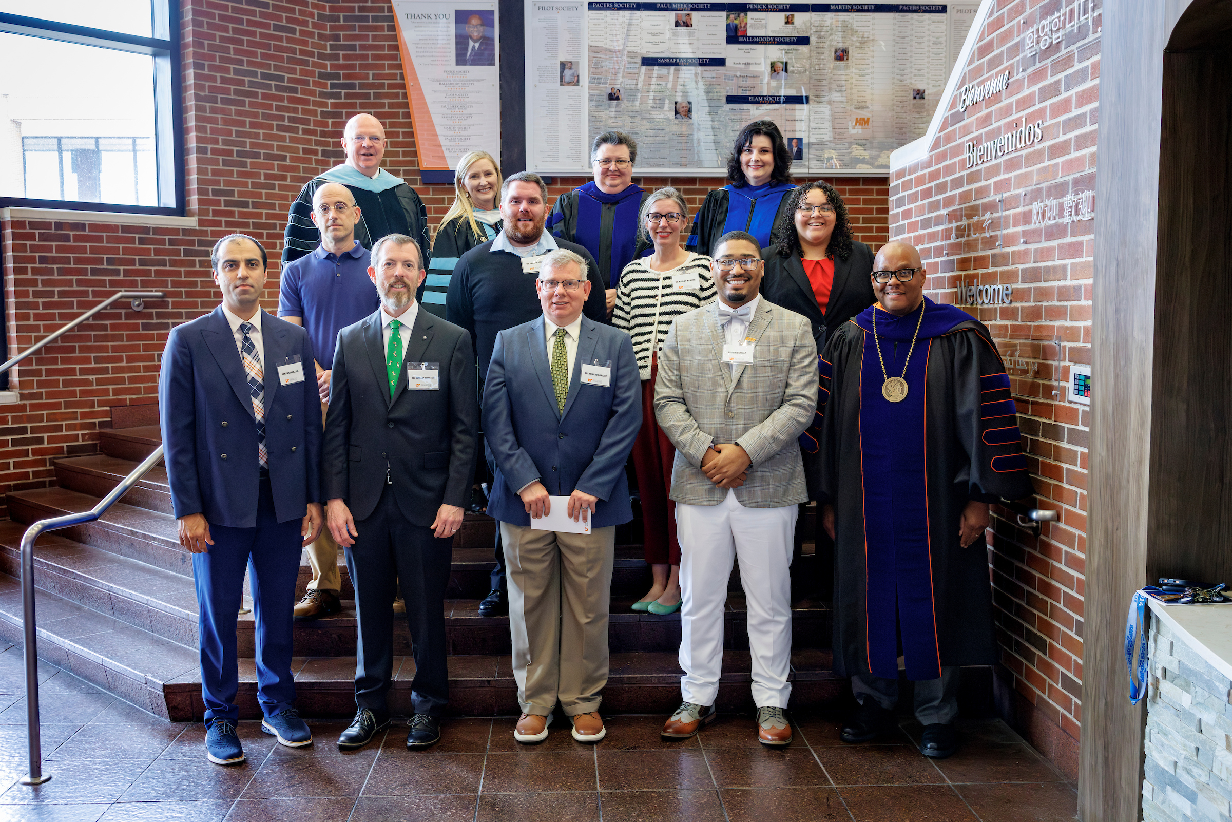 A group of people standing on an indoor staircase with a brick wall and plaques in the background.