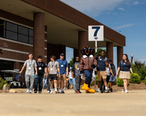 A group of people walking with a bird mascot near a brick building. A person holds a sign with the number 7.
