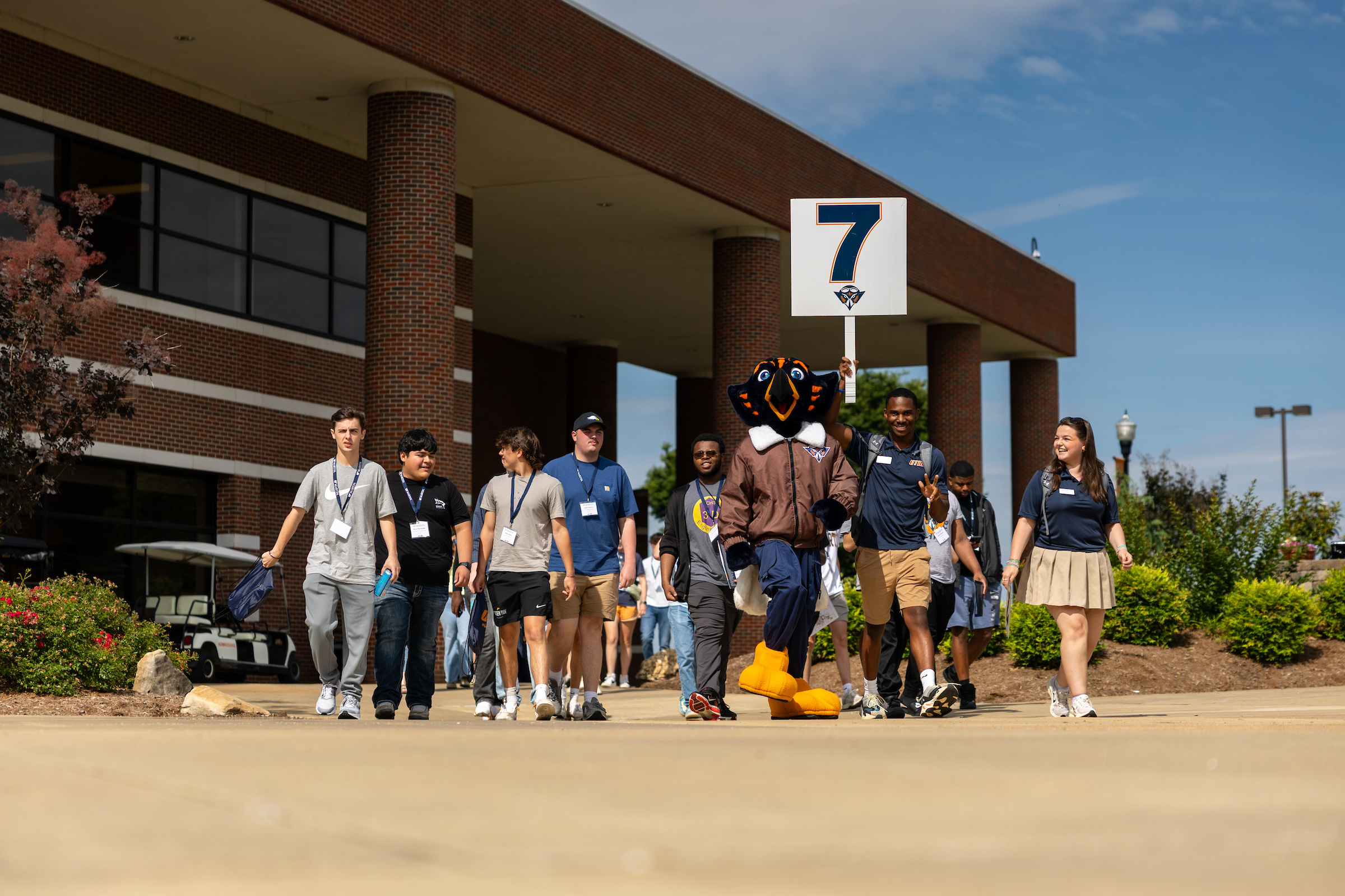 A group of people walking with a bird mascot near a brick building. A person holds a sign with the number 7.