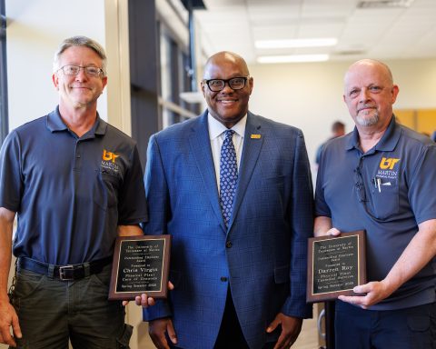 Three men standing indoors, the outer two holding plaques.