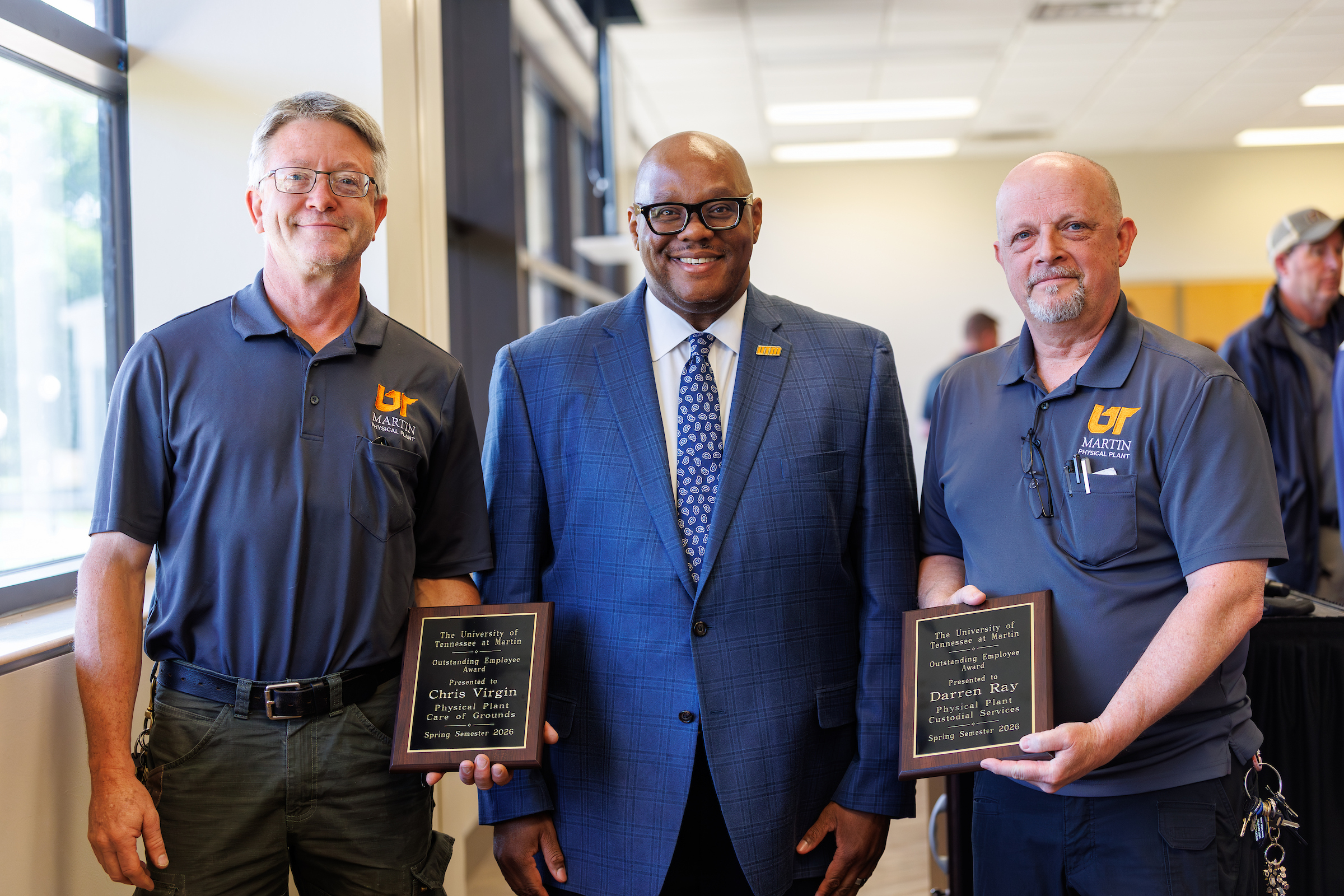 Three men standing indoors, the outer two holding plaques.