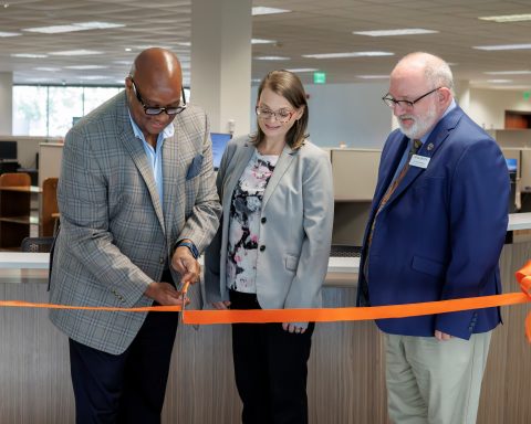 Three people in an office setting at a ribbon-cutting ceremony with an orange ribbon.