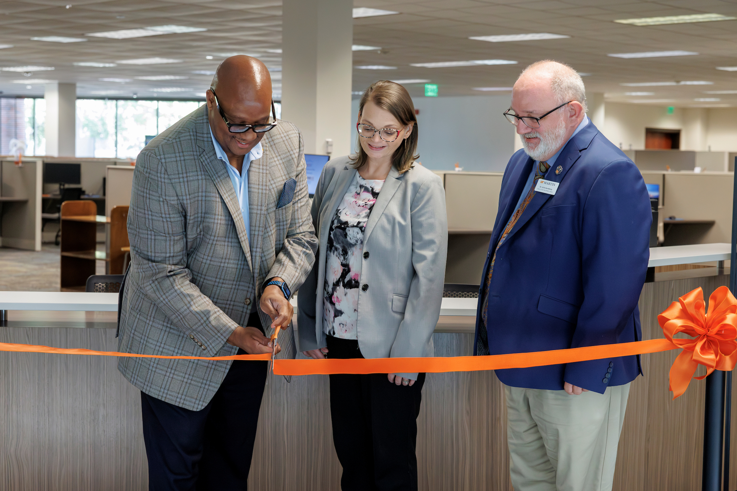 Three people in an office setting at a ribbon-cutting ceremony with an orange ribbon.