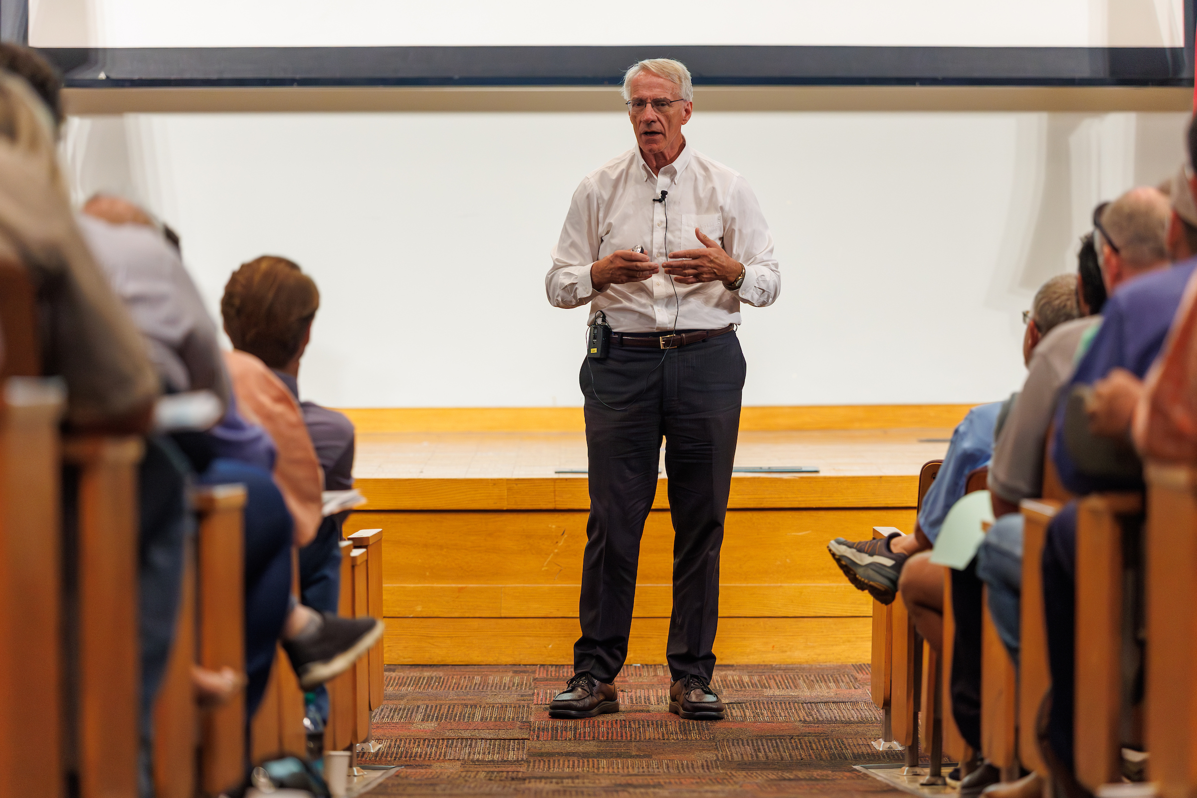 A man is standing in front of a stage in an auditorium speaking to the people seated in the audience.