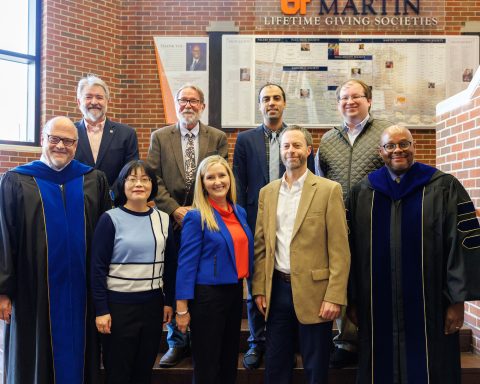 A group of nine diverse individuals poses on a staircase, with some wearing academic regalia, in a brick interior with a giving societies display in the background.