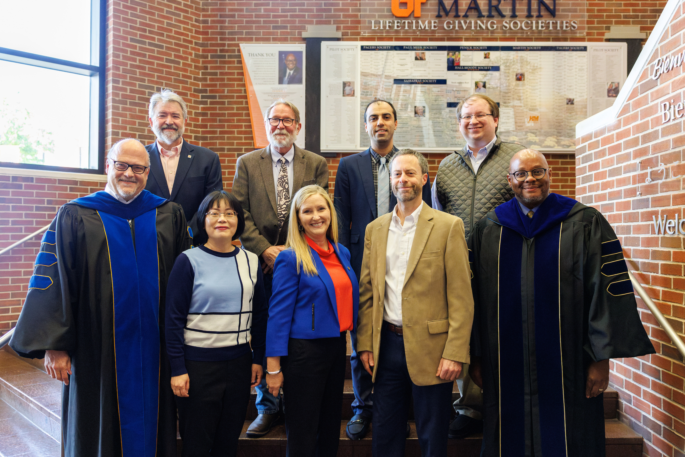A group of nine diverse individuals poses on a staircase, with some wearing academic regalia, in a brick interior with a giving societies display in the background.