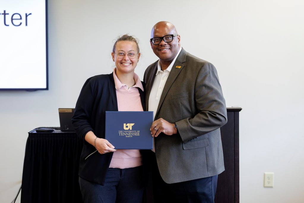 Two individuals stand together indoors, smiling while holding a dark blue folder with the University of Tennessee logo. One wears glasses and a black blazer, while the other is in a gray blazer.