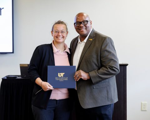 Two individuals stand together indoors, smiling while holding a dark blue folder with the University of Tennessee logo. One wears glasses and a black blazer, while the other is in a gray blazer.