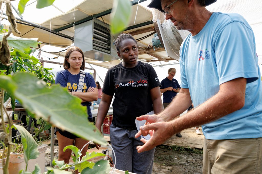 A greenhouse scene with two women listening to a male instructor explaining plant care. One woman looks contemplative while the other appears engaged.