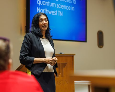 A woman in a black blazer speaks confidently in front of a blue presentation screen about quantum science, with blurred audience members in the foreground.