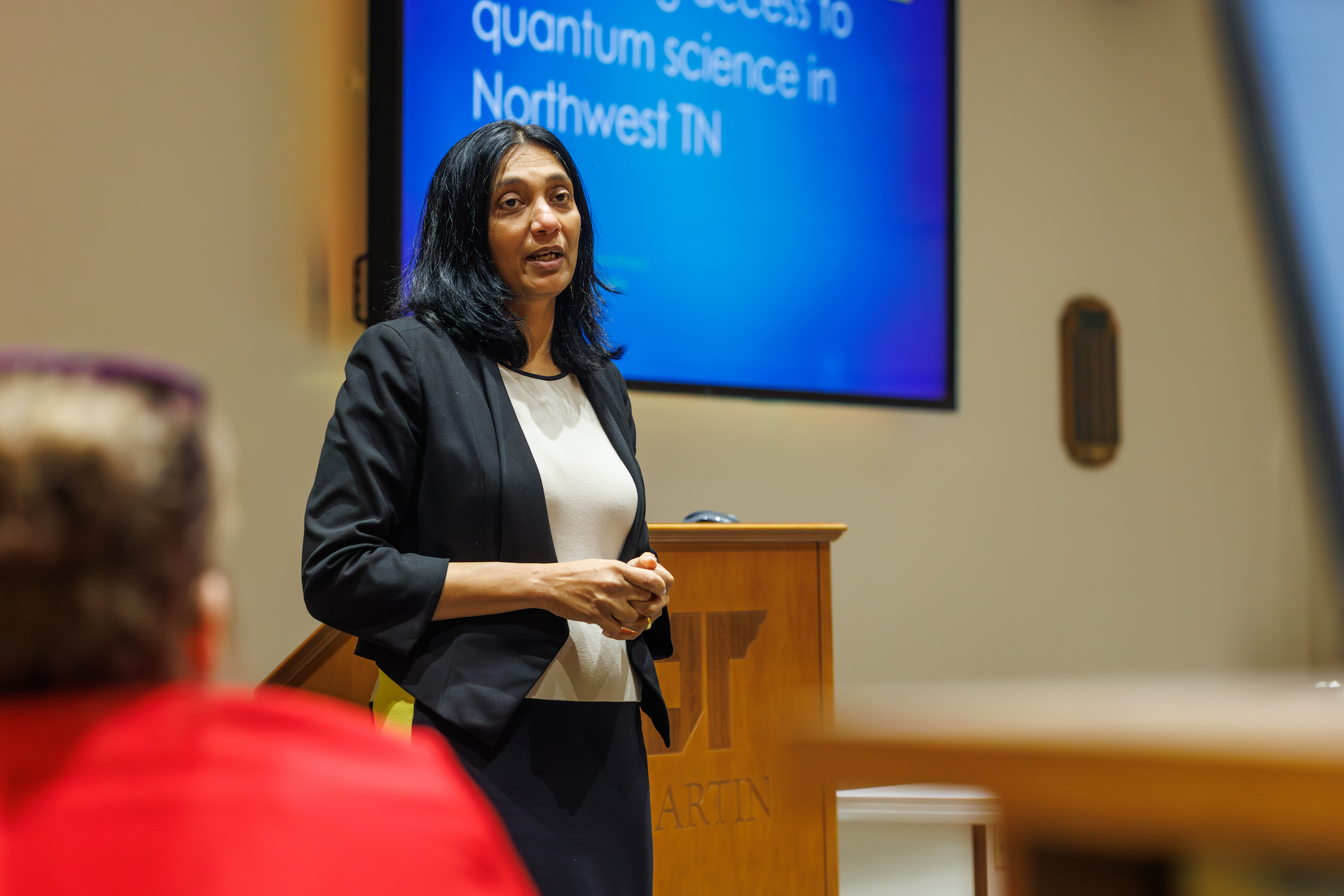 A woman in a black blazer speaks confidently in front of a blue presentation screen about quantum science, with blurred audience members in the foreground.