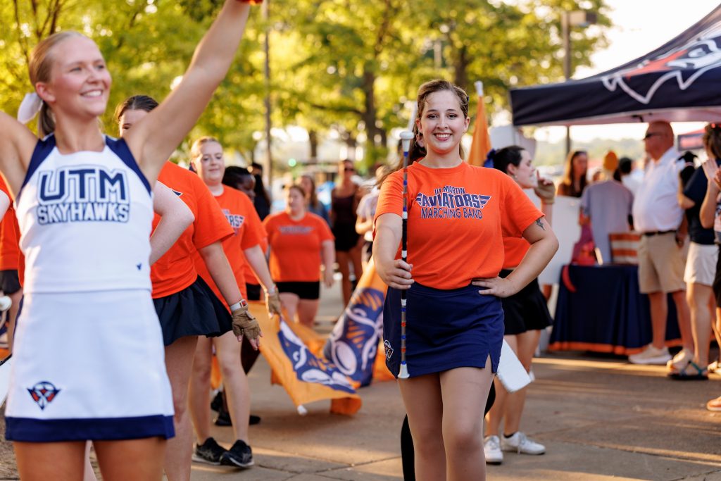 A cheerful cheerleader and a smiling band member hold their positions in a marching band parade outdoors, surrounded by fellow band members in orange t-shirts and the backdrop of green trees.