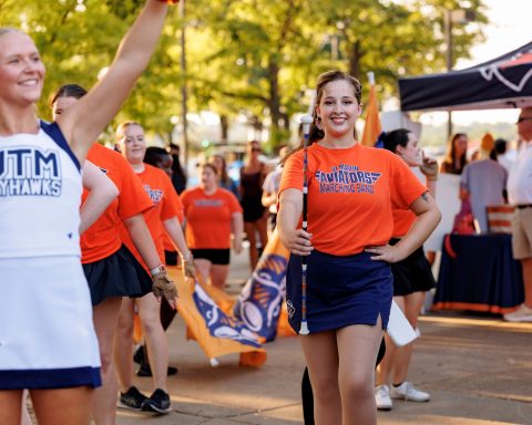 A cheerful cheerleader and a smiling band member hold their positions in a marching band parade outdoors, surrounded by fellow band members in orange t-shirts and the backdrop of green trees.