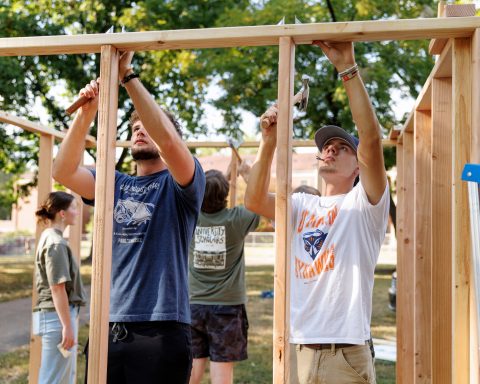 A group of young adults works together to build a wooden structure outdoors, with two men using hammers and a woman observing in the background.