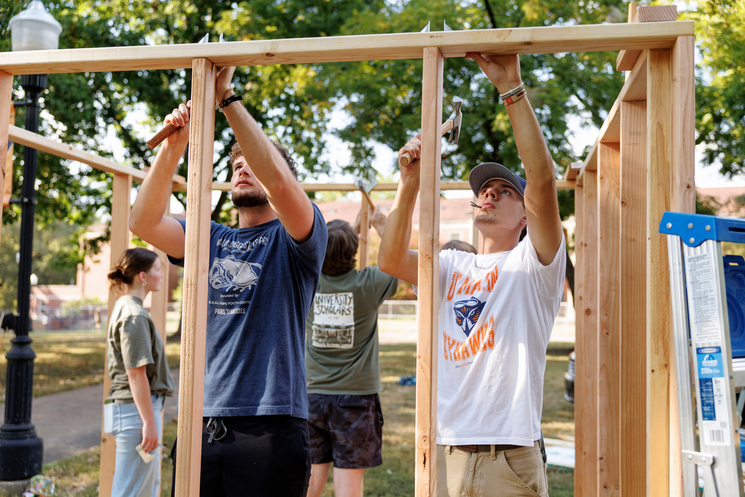 A group of young adults works together to build a wooden structure outdoors, with two men using hammers and a woman observing in the background.