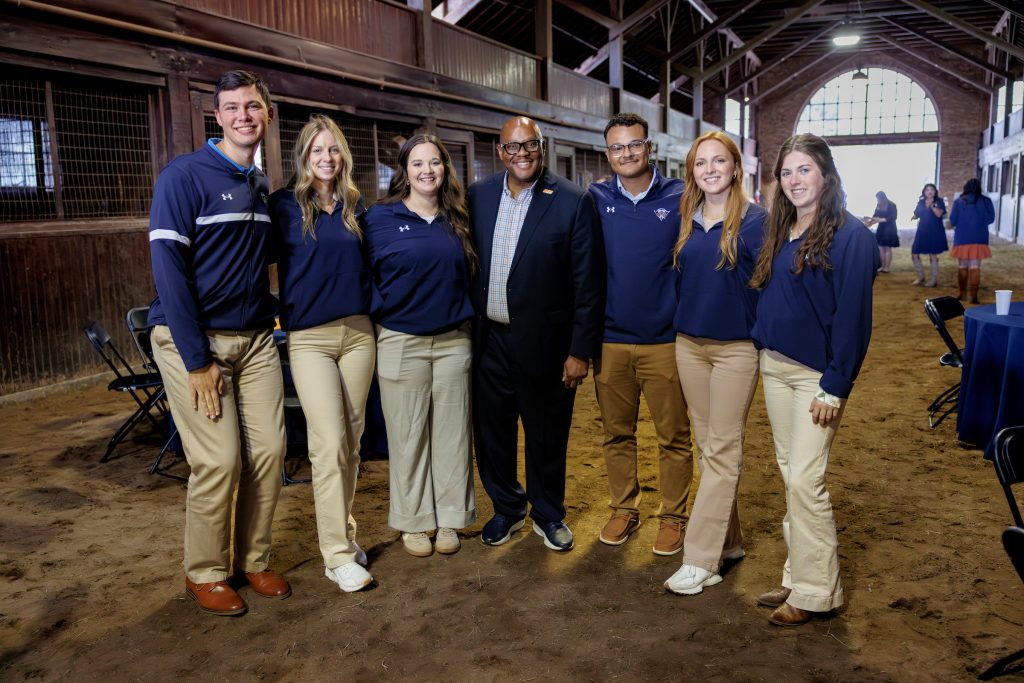 A group of six people, wearing matching navy blue shirts and light pants, pose together in a rustic barn setting. The central figure, in a suit jacket, stands surrounded by three women and two men, all smiling.