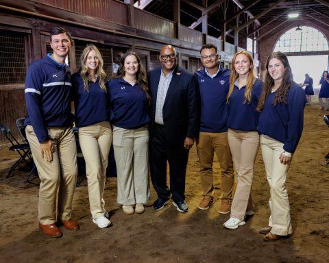 A group of six people, wearing matching navy blue shirts and light pants, pose together in a rustic barn setting. The central figure, in a suit jacket, stands surrounded by three women and two men, all smiling.
