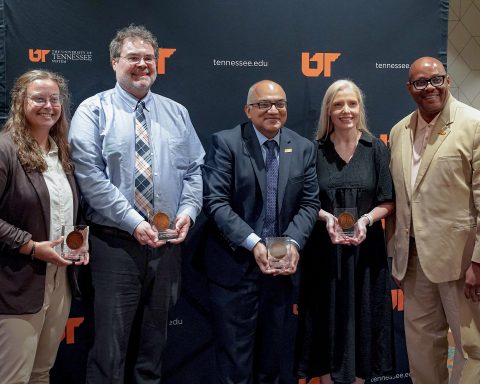 Four individuals holding awards stand together with a man in a tan suit in front of a dark University of Tennessee backdrop during a formal event.
