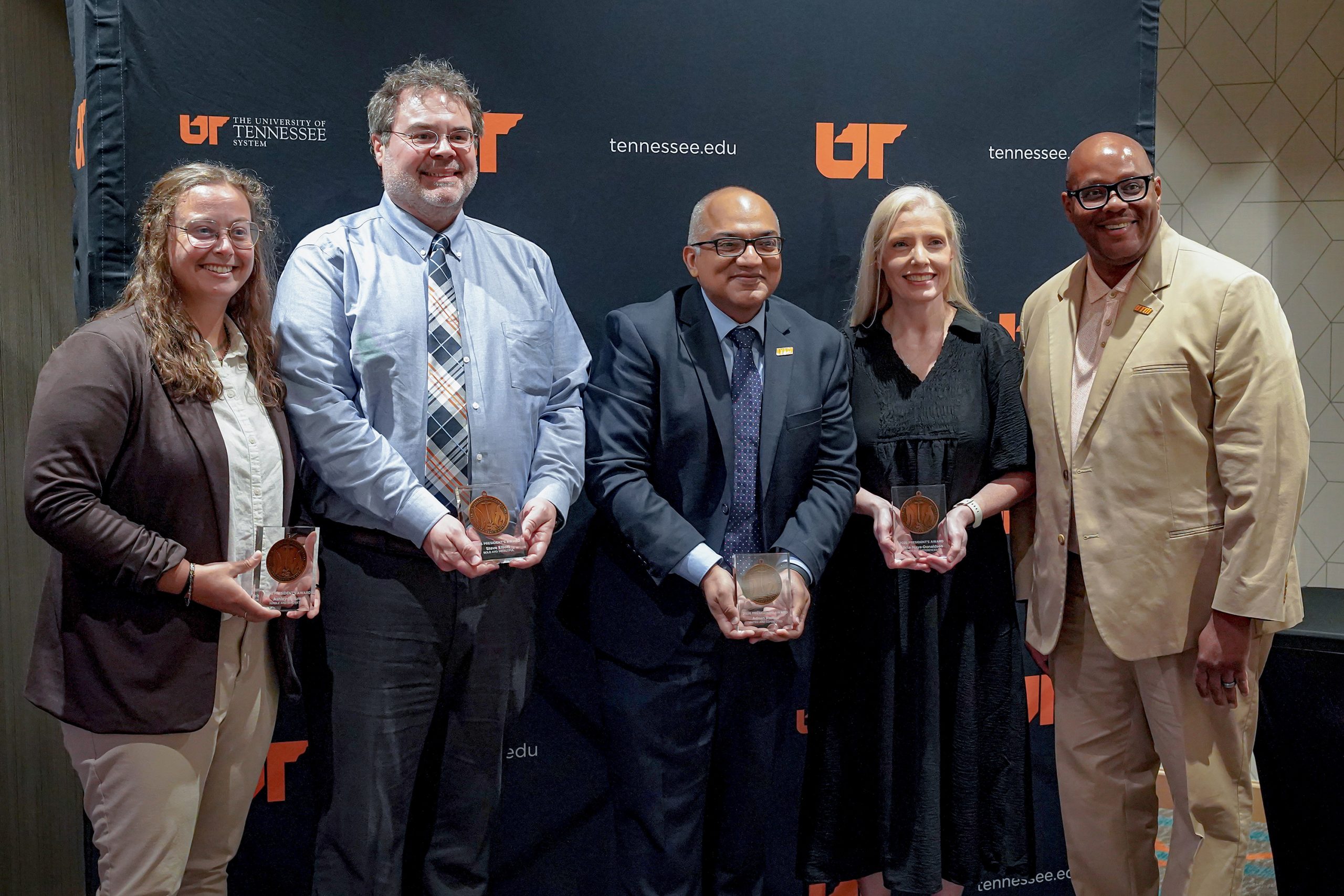 Four individuals holding awards stand together with a man in a tan suit in front of a dark University of Tennessee backdrop during a formal event.