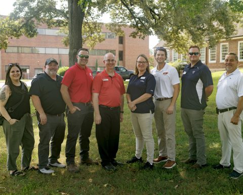 Eight people are posed in a tree-shaded area of a campus