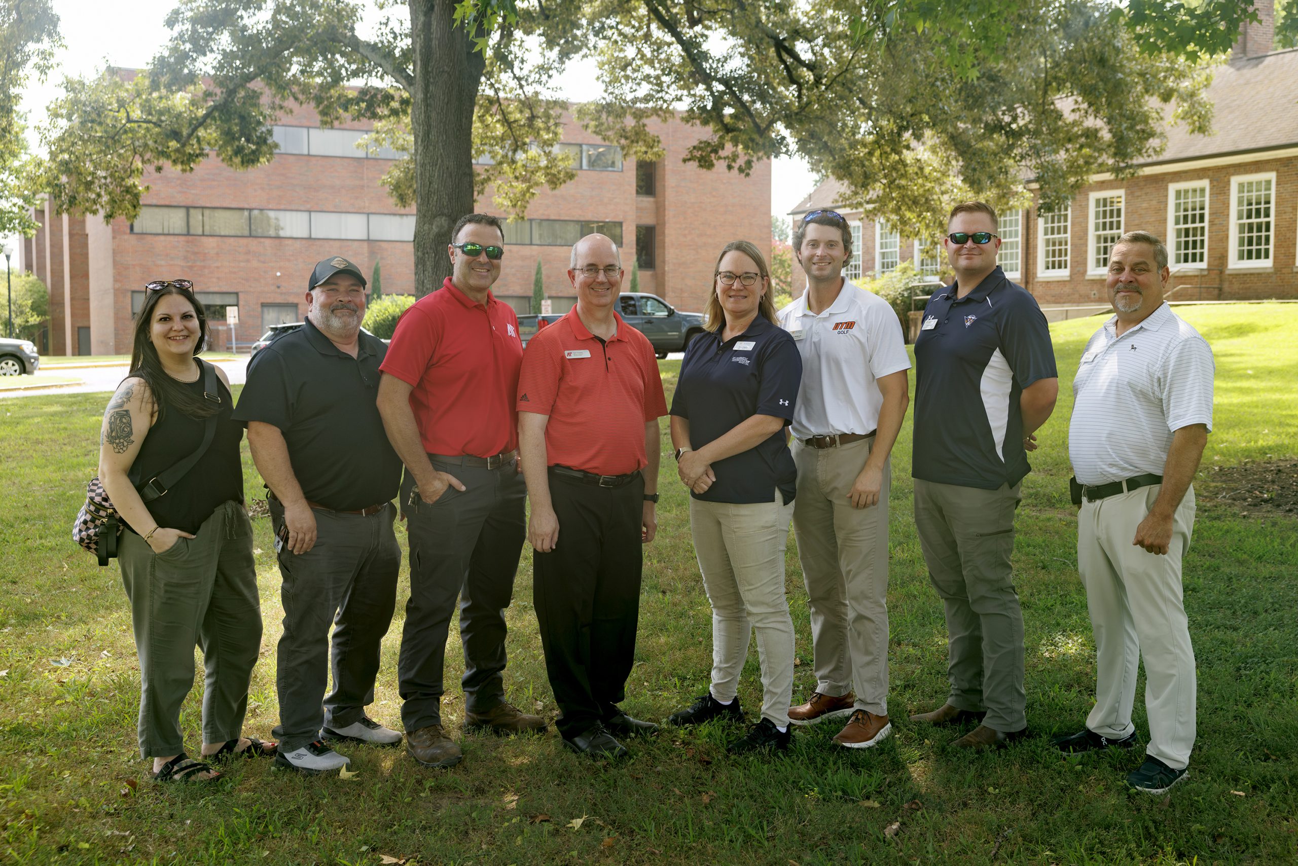 Eight people are posed in a tree-shaded area of a campus