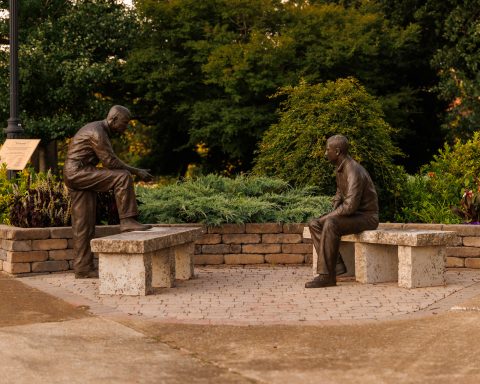 Statues of two men, one seated and one standing with his foot resting on a bench, are shown