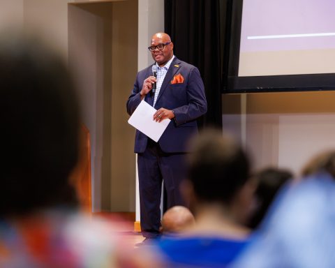 A man in a navy suit with an orange pocket square speaks into a microphone while holding papers at the front of a meeting room. Audience members are visible in the foreground, and a projection screen hangs behind him.