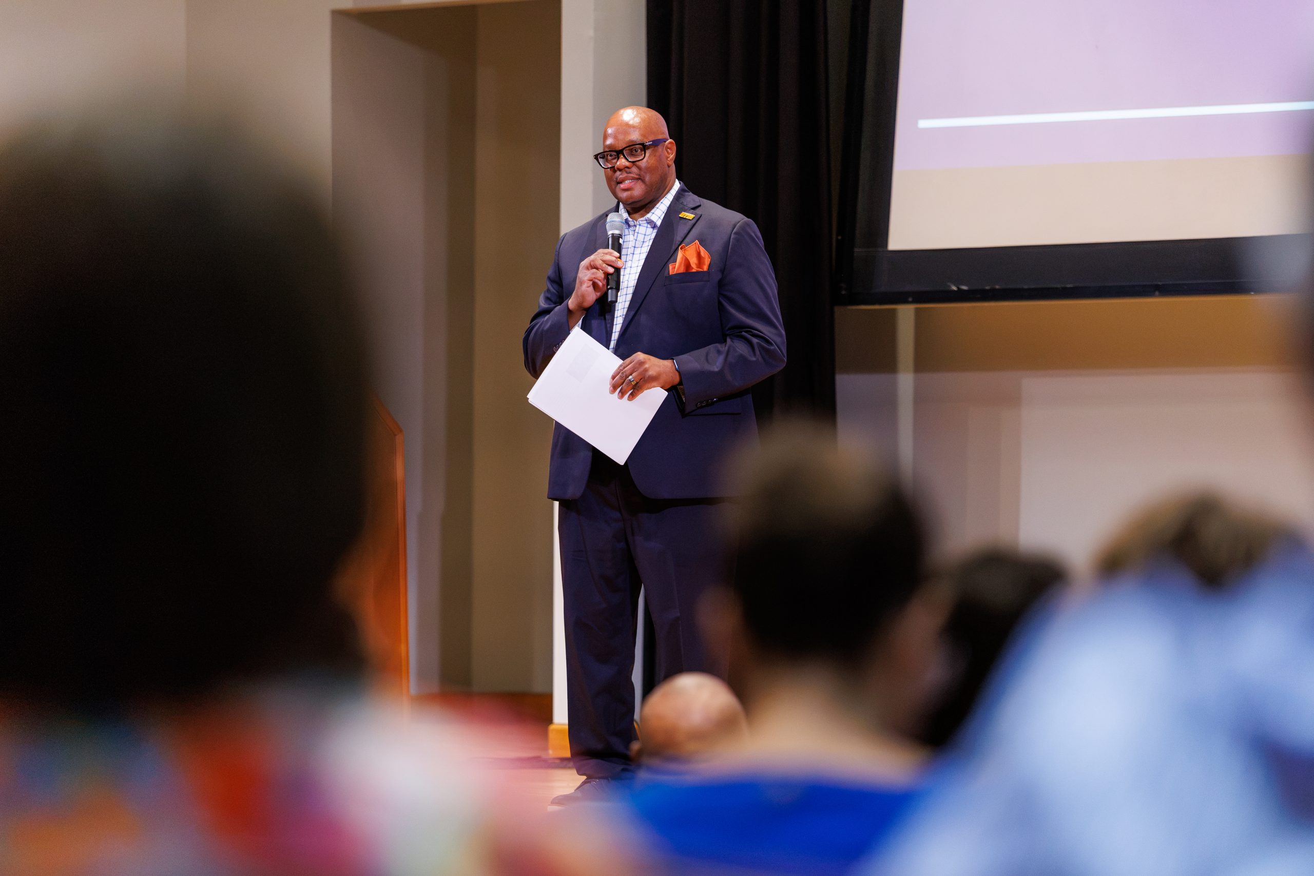 A man in a navy suit with an orange pocket square speaks into a microphone while holding papers at the front of a meeting room. Audience members are visible in the foreground, and a projection screen hangs behind him.