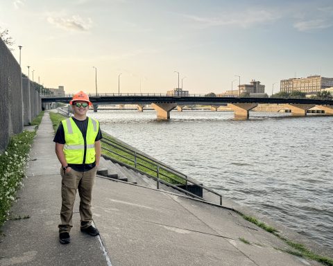 A person wearing an orange hard hat, reflective safety vest, sunglasses and work pants stands along a paved riverfront walkway. A bridge crosses the river behind them, with several downtown buildings visible across the water in the distance.