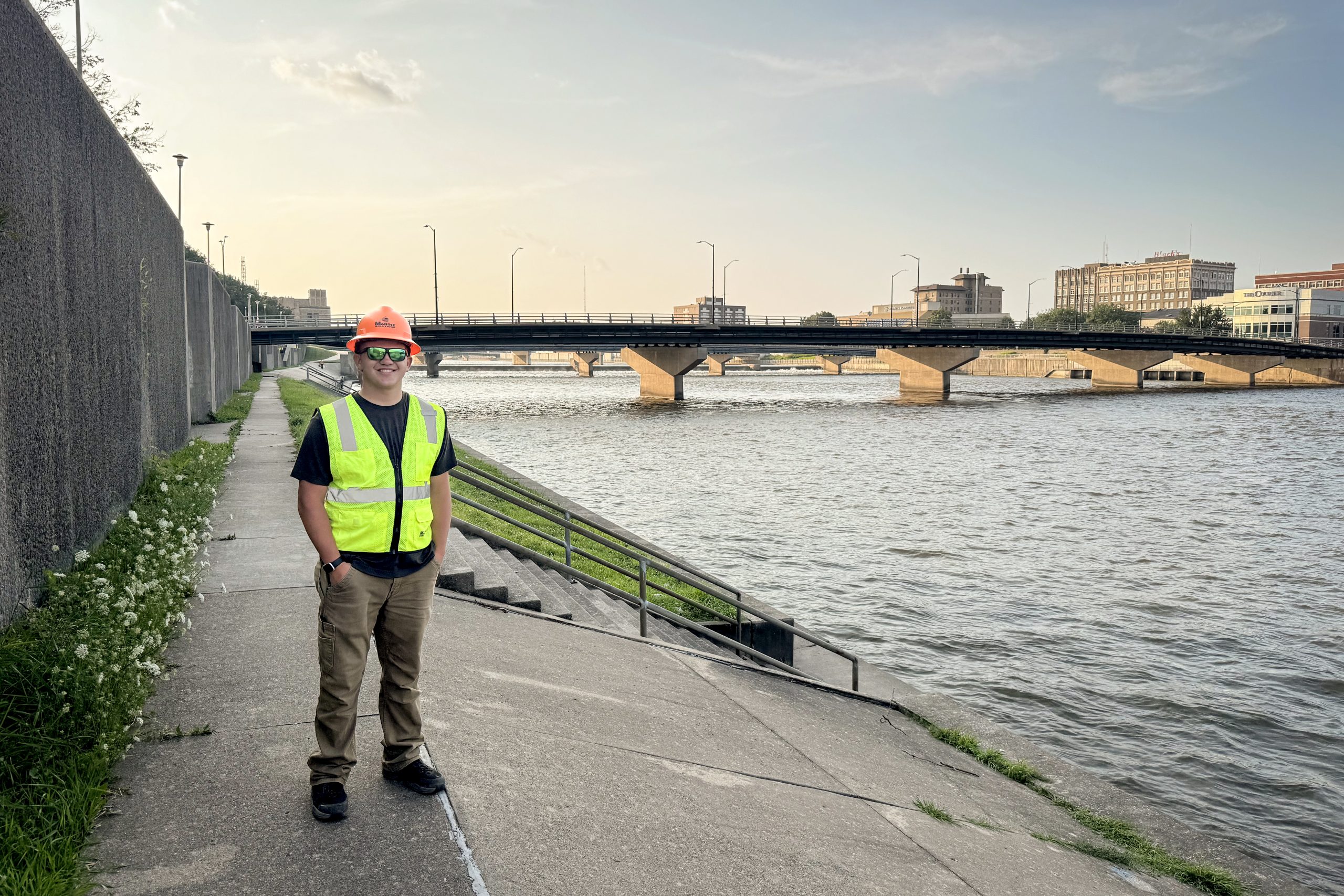 A person wearing an orange hard hat, reflective safety vest, sunglasses and work pants stands along a paved riverfront walkway. A bridge crosses the river behind them, with several downtown buildings visible across the water in the distance.