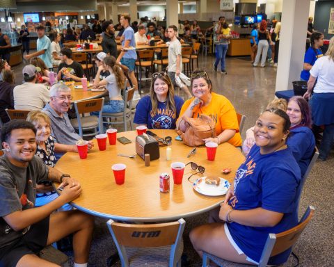 A group of UT Martin students and guests smile for the camera while seated around a table during a crowded welcome-back picnic in the university dining hall. Other attendees gather, eat and socialize throughout the room.