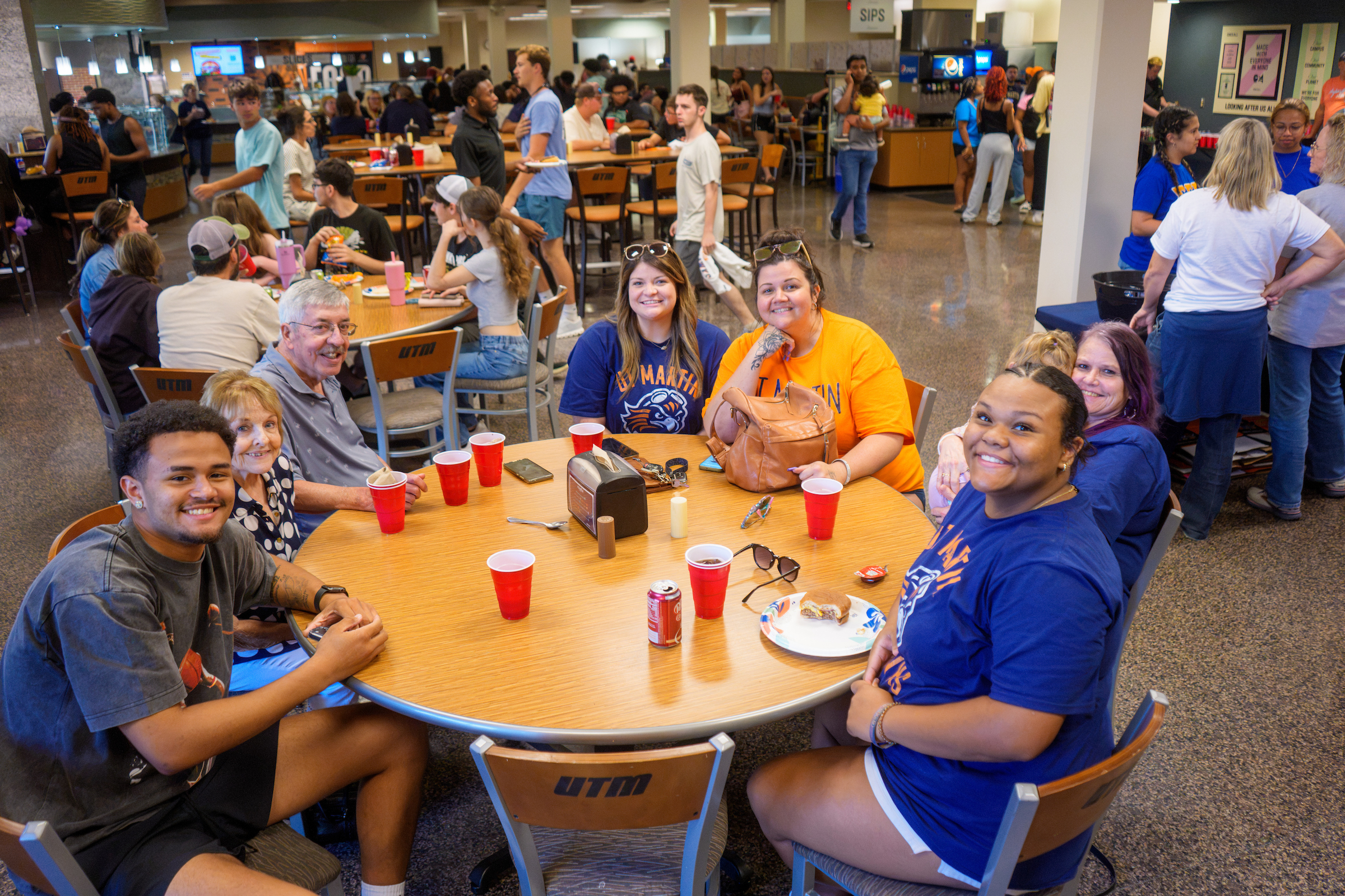 A group of UT Martin students and guests smile for the camera while seated around a table during a crowded welcome-back picnic in the university dining hall. Other attendees gather, eat and socialize throughout the room.