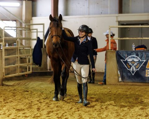 A young lady dressed in horse riding clothes smiles as she walks with a horse