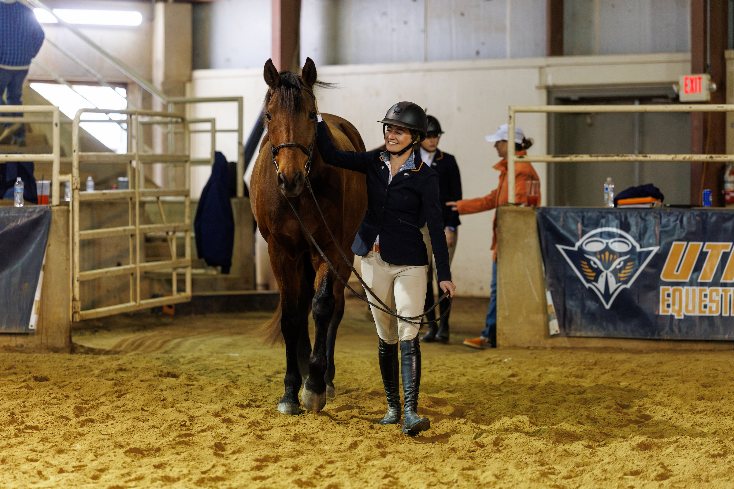 A young lady dressed in horse riding clothes smiles as she walks with a horse