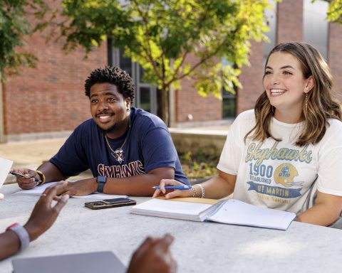 Two UT Martin students smile while studying with friends at an outdoor table on campus.