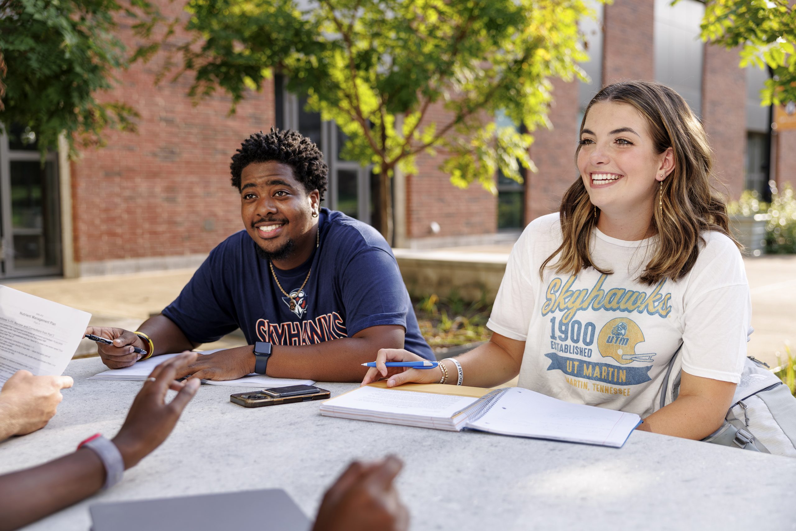 Two UT Martin students smile while studying with friends at an outdoor table on campus.
